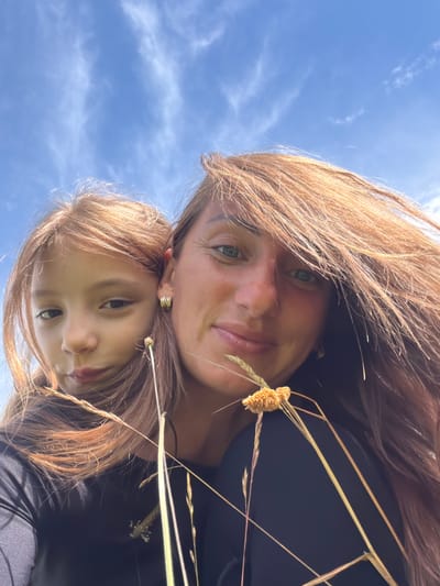This is a close-up selfie taken outdoors, with a bright blue sky and scattered white clouds in the background. Two individuals, an adult and a child, are featured. The adult, positioned on the right, has long, reddish-brown hair blowing in the wind. They are wearing a black top and have blue-green eyes and a visible earring. The child, on the left, has similar reddish-brown hair and is looking towards the camera with a neutral expression. They are also wearing a black top. In the foreground, there are several dry stalks of grass and a small, fuzzy yellow flower-like object, suggesting an outdoor, possibly rural or park-like setting. The lighting indicates it is daytime. No specific landmarks are visible to pinpoint a location within Jēkabpils, Latvia.