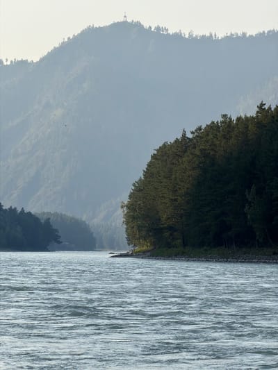 The image depicts a wide, choppy river flowing through a mountainous, forested landscape. In the foreground, the river's surface is covered in numerous ripples and waves, reflecting the hazy daylight. The riverbanks are lined with dense, dark green coniferous trees, creating a sharp contrast with the muted tones of the distant mountains. On the right bank, a rocky shore is visible, with a few small figures, likely people, standing near the treeline. A small group of trees, possibly birch, with lighter bark, are interspersed among the darker conifers.
In the background, a large, hazy mountain range dominates the scene. The slopes of the mountains are covered in vegetation, and the summit is shrouded in a light mist. A man-made structure, resembling a cross or a small monument, is visible at the very peak of the mountain, silhouetted against the bright sky. The overall atmosphere is serene and natural, with a hint of human presence.
The scene is located at the Katun River near the village of Anos in Russia. The Katun River is a major river in the Altai Republic and Altai Krai, flowing through a region known for its stunning natural beauty and mountainous terrain. The village of Anos itself is situated on the banks of the Katun River, surrounded by picturesque landscapes.