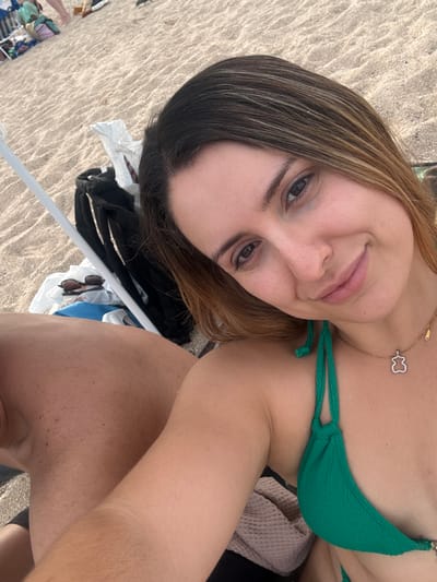 A woman with light brown hair, wearing a green bikini and a necklace with a small bear pendant, takes a selfie on a sandy beach in Vilassar de Mar, Spain. She is smiling at the camera, with her eyes looking slightly to the side. Behind her, a person's back and shoulder are visible, as well as a white beach umbrella, a black bag, and sunglasses. The background consists of the textured sand of the beach, with faint footprints and some other beachgoers visible in the distance. The sea is not visible in the frame.