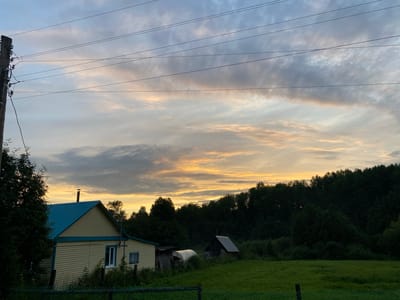 This media file captures a rural scene in Baklushi, Russia, likely during either sunrise or sunset. The sky is filled with dramatic clouds, tinged with hues of orange and yellow from the sun, contrasting with the cooler blues and grays. Power lines stretch across the sky, supported by a weathered utility pole on the left side of the frame.
In the foreground, a light yellow house with a bright blue roof is visible. It has at least two windows and a chimney. Adjacent to the house, a small greenhouse structure and a dark shed are present. Further to the right, a smaller, darker wooden structure stands in a grassy field. The landscape is bordered by a dense line of dark green trees that create a stark silhouette against the sky. The overall impression is one of tranquility and the peacefulness of the countryside. There is no visible text in the image.