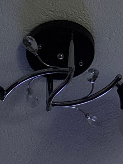 The image shows a close-up, low-angle view of a modern, black ceiling light fixture with chrome accents and clear crystal-like elements. The fixture has a circular black base plate with several screws visible. From the base plate, chrome arms curve outwards, each terminating in a point or adorned with a faceted crystal. One arm on the left has a crystal hanging from it, and another on the right has a similar crystal. The crystals appear to be teardrop-shaped or multifaceted. The light fixture is mounted on a textured, light-colored ceiling, which has a slight blueish hue due to the lighting. The overall impression is one of contemporary interior design. No people or actions are visible in the frame. The image is a still photograph and does not provide any information about the city of Gurkovo, Bulgaria, or its context within that location.