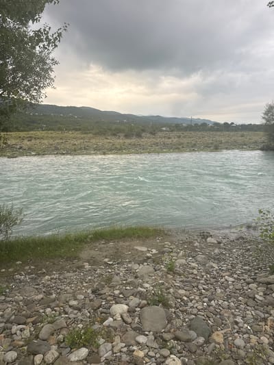 The image depicts a wide, turquoise river flowing through a valley. The foreground is a rocky shore with scattered green vegetation. The river's surface is rippled, indicating movement. Across the river, a vegetated hillside with scattered houses can be seen, leading up to rolling hills and mountains in the background. The sky is overcast with a mix of gray clouds and patches of lighter sky, suggesting diffused daylight. The scene appears to be in a natural, rural environment. The location is specified as Aragvispiri, Georgia. There are no people or animals visible in the image. The overall atmosphere is calm and natural.