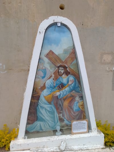 A mounted religious image depicting Jesus carrying the cross, supported by a woman, is affixed to a tan-colored wall with a circular hole. The image is framed in white and is arched at the top. The artwork includes three figures: Jesus, the woman, and a figure behind Jesus in the background with a helmet on. There is a small plaque below the painting. The scene is at the location in the city of Municipio Tarija, Bolivia. Yellow plants are growing at the base of the image.