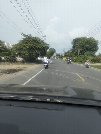 The image captures a street scene in Payameuneng, Indonesia, on an overcast day. A paved road with white and yellow lane markings dominates the foreground and middle ground. Several motorcycles are in motion. On the right side of the road, a person on a red and white motorcycle travels away from the viewer. Further down the road, another person on a similar motorcycle is also heading away. On the left side of the road, a person wearing a white outfit on a white scooter is moving towards the viewer. In the background, lush green trees line the sides of the road, interspersed with utility poles and power lines. Buildings and other structures are visible beyond the trees on the left. The perspective is from inside a vehicle, with the dashboard and windshield wiper visible at the very bottom of the frame. The overall impression is one of a typical day on a road in a semi-urban or rural Indonesian setting.