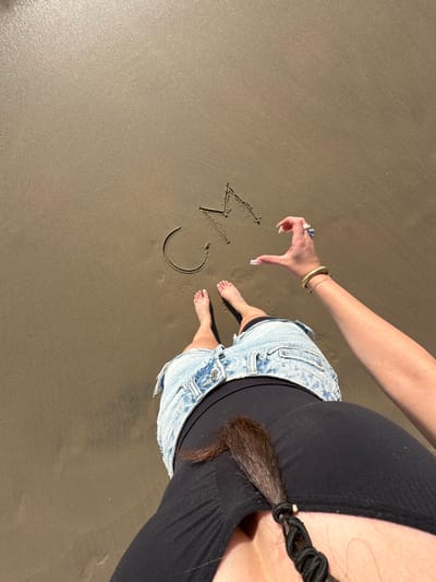 A high-angle forensic view captures a person standing on wet sand at Manhattan Beach, United States. The individual's bare feet are visible, positioned on a smooth, damp sandy surface. The person wears light blue denim shorts or a skirt and a dark top. A dark brown braided or twisted section of hair hangs forward from the upper torso area. The person's right arm extends into the frame, with the hand positioned above the sand, forming the right half of a heart shape with its fingers. The hand displays long, light-colored fingernails, a ring on the ring finger, and another on the middle finger. The right wrist is adorned with a ribbed gold-colored cuff bracelet and a thinner gold-colored chain bracelet. On the sand, the initials "CM" are distinctly etched to the left of the hand.