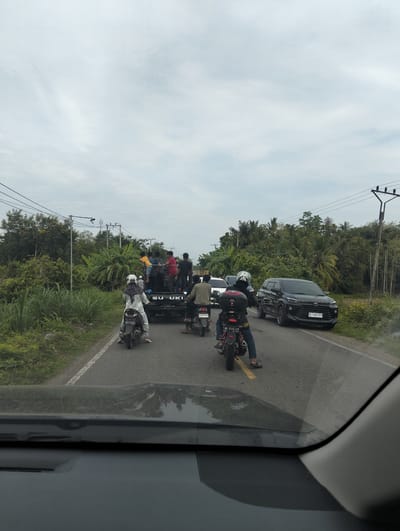 In Tanjongkeumala, Indonesia, a traffic jam unfolds on a two-lane asphalt road lined with lush vegetation. A black Suzuki pickup truck, loaded with several passengers including children, leads the procession. Following closely are three motorcycles. The first motorcycle, ridden by a person in a white helmet and patterned headscarf, is partially obscured by the pickup. The second motorcycle, a black scooter, carries a rider in a black helmet and jacket. The third motorcycle is a smaller, red and black scooter with a rider in blue jeans and a black helmet. On the right side of the road, a black Toyota Avanza is parked. The sky is overcast, and power lines stretch across the scene, supported by utility poles. The view is captured from inside a vehicle, with the dashboard and windshield visible in the foreground.