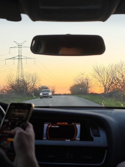 The image depicts a view from inside a car traveling on a road near Gorni Tsibar, Bulgaria, facing towards the horizon at sunset. The dashboard and interior of the car are visible, including a hand holding a mobile phone and the car's infotainment display. The car is moving along a straight road with a car approaching from the opposite direction. Power lines and utility poles are visible in the background, flanking either side of the road, as are trees without leaves. The car's rearview mirror is visible.