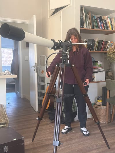 A person is operating a telescope indoors. The telescope is white with a black lens cover, mounted on a brown wooden tripod. The person, wearing a burgundy fleece jacket, black pants, and socks with sandals, is adjusting the telescope's controls. The room contains a bookshelf filled with books, a basket, a stepladder, and other furniture. A window is visible in the background. The specific location in La Lucila, Argentina, cannot be determined from the image.