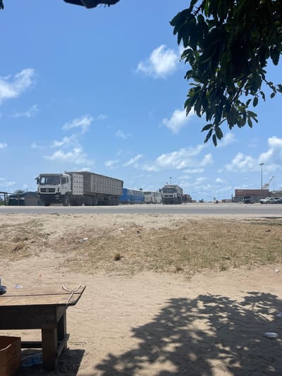 The scene depicts a sunny daytime view in Ebute Lekki, Nigeria, characterized by an industrial or transport environment. The sky is clear blue with scattered white cumulus clouds.

In the foreground, an unpaved sandy expanse with sparse, dry grass occupies the lower half of the frame. Scattered small pieces of white debris are visible on the sand. On the far left, partially obscured by shadow, is a dark wooden table with a coiled white string or rope and a small white object on its surface. A brown cardboard box rests beneath the table. Dark, irregular shadows from an overhead tree are cast upon the sandy ground, primarily on the right side.

A paved road crosses the midground horizontally. On the left side of the road, a large white multi-axle dump truck, loaded with light-colored material, is prominently positioned, facing slightly towards the left. To the right of this truck, a line of several blue and white buses is parked. Further right along the road, another white truck is visible.

The background reveals elements of an industrial or port facility. A red shipping container is visible on the far right, alongside a white vehicle and a tall industrial crane (possibly a gantry or container crane). Various structures and utility poles are distributed across the distant horizon.

A large, dark green leafy branch from an unseen tree extends into the upper right corner of the frame, partially obscuring the sky. No individuals are present in the image, and no discernible actions or interactions are occurring; the vehicles appear stationary.