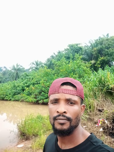 A man wearing a red baseball cap and a black t-shirt is pictured in a close-up selfie. He has a beard and is looking directly at the camera. The background features a murky body of water to the left, with a dense green forest and tall grass lining its edge. The sky is overcast and bright white. The overall scene appears to be a natural setting, possibly near a river or stream in the city of Nanka, Nigeria. The ground in the foreground is composed of dirt and dry grass with some scattered debris.