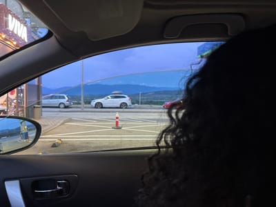 The image, taken from inside a vehicle, shows a scene at dusk. The view is through the car's side window and the windshield. In the foreground, a person's dark, curly hair obscures the right side of the frame. The interior of the car is visible, including the door panel and handle on the left.
Outside the car, a gas station with pumps is reflected in the driver's side mirror. Beyond the gas station, a silver station wagon is parked. Further in the background, a white SUV with a roof rack is positioned on a roadway. A red car is also visible. The landscape features hills or mountains in the distance under a twilight sky with shades of blue and purple. White lines mark lanes on the asphalt. A short orange and white traffic cone stands on the road. A trash can is next to the silver car. The scene appears to be a roadside service area or a gas station on the outskirts of Kalugerovo, Bulgaria, given the presence of a "VIVA" sign, possibly indicating a chain or brand associated with such services in the region.