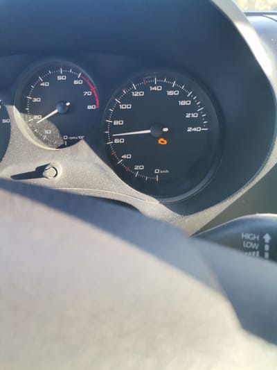 The image displays a close-up view of a car's dashboard, specifically the instrument cluster, from the driver's perspective, likely captured during daylight hours in Batovo, Bulgaria. The interior surfaces of the dashboard and instrument panel show a noticeable layer of dust.
The left gauge is a tachometer, indicating the engine is running at approximately 2000-2500 RPM (reading between '20' and '30' on the 'rpm x 100' scale). The right gauge is a speedometer, showing the vehicle is currently traveling at approximately 80 km/h. The speedometer's maximum reading is 240 km/h.
A prominent detail is the illuminated orange "Check Engine" warning light visible beneath the speedometer needle, indicating a potential issue. Part of a control stalk is visible on the right, featuring text "HIGH" and "LOW" and an upward arrow, likely controlling headlights or wipers. A small adjustment knob or button is located on the lower left of the instrument cluster. The bright sunlight creates reflections and shadows across the dashboard.
Visible text includes "rpm x 100", numerical values "10" through "80" on the tachometer, and "km/h" with numerical values "20" through "240-" on the speedometer. The control stalk also clearly displays "HIGH" and "LOW".