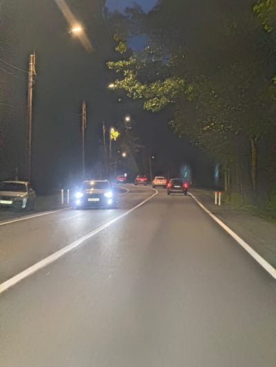 A nighttime, eye-level, medium shot captures a road in Ushkovo, Russia, illuminated by streetlights and vehicle headlights. Several cars are visible driving on the asphalt road, which has a white line marking its edge. On the left side of the road, a single, tall wooden utility pole stands, with electrical wires extending from it. Adjacent to the road on the left, a silver car is parked. Dense trees line the right side of the road, with some foliage extending over the asphalt. Two white road markers with red tops are positioned near the edge of the road on the right. The sky is dark, indicating the late hour.