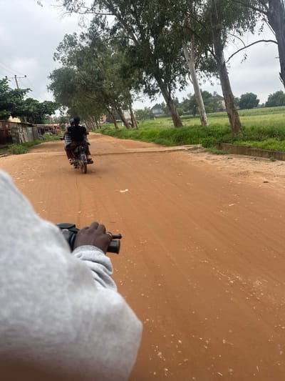 An image taken from the perspective of someone riding a motorcycle in Katambe, Nigeria. The motorcycle is traveling on a dirt road. In front of the motorcycle, another motorcycle is visible, carrying two people. The road is lined with trees on both sides and some buildings are visible in the distance. The sky is overcast.