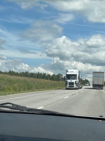 The image depicts a view from inside a vehicle, looking out at a highway on a partly cloudy day. The camera is angled, showing the road curving away to the right. In the foreground, a windshield wiper is visible.

On the highway, there are several trucks. The closest truck is a white cab-over tractor unit, facing away from the viewer. Further down the road, a white semi-trailer truck with a tan trailer is visible. The trailer has Russian text on it, including "ПРИГЛАШАЕМ ЗАРАБОТАТЬ" and "8-800-246-015". There's also a smaller white vehicle, possibly a car, behind the semi-trailer truck.

The roadside to the left of the highway is a grassy field with scattered wildflowers and a line of trees in the distance. The sky is a bright blue with large, fluffy white and gray clouds, suggesting fair weather. The lighting indicates it is daytime, likely afternoon. The location is identified as Bavly, Russia.