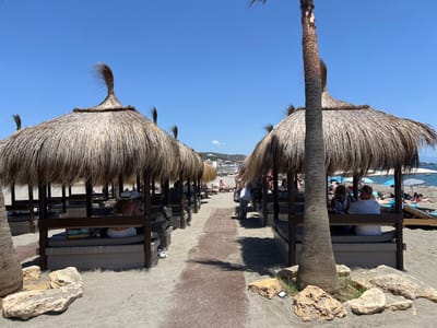 The image depicts a sandy beach in San Roque, Spain, on a sunny day with a clear blue sky. The foreground is dominated by rows of thatched-roof cabanas offering shade to beachgoers. These cabanas are furnished with cushioned benches. Several people are visible sitting or lounging within these structures. In the middle ground, a pathway runs between the cabanas, leading towards the sea. Beyond the cabanas, the beach continues with more lounge chairs and umbrellas. The background shows the Mediterranean Sea with a few boats and a distant town nestled on a hill. The overall scene conveys a relaxed and leisurely atmosphere at a popular beach resort.