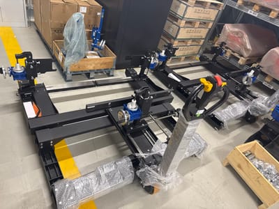 The image depicts an indoor industrial facility, identified as being in Logatec, Slovenia. The concrete floor is marked with yellow safety lines. The primary subjects are multiple newly manufactured industrial machines, characterized by black rectangular frames, horizontal rails, blue-colored motorized components, and metallic shafts on adjustable mounts.

In the foreground, an electric pallet jack, primarily black with a yellow and red control handle, is visible. Its lower structure and handle are completely encased in clear protective plastic film. Adjacent to the pallet jack on the right, a wooden crate contains numerous small metallic components.

In the background, stacked brown cardboard boxes rest on a wooden pallet. One wooden crate on this pallet holds a blue component extensively wrapped in clear bubble wrap. To the right, a multi-tiered metal shelving unit is stocked with additional wooden pallets, loaded with items covered in red and clear plastic sheeting. Several shelves are marked with labels, including "01-413" and "01-382." No individuals are present in the scene.