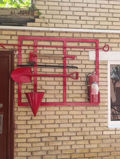 A red metal rack is mounted on a light brown brick wall, holding firefighting equipment. The rack contains an axe, two shovels, a bucket, and a fire extinguisher. The axe is at the top left, followed by a black rod with a red handle, then a red shovel, and finally a red bucket hanging below. To the right of the bucket, a red fire extinguisher is mounted in a bracket. The wall is made of light brown, rectangular bricks laid in a stretcher bond pattern. Above the rack, a section of a white window frame is visible, and green vines grow from the top left corner of the image, trailing down the wall. A dark brown door with a silver handle is on the left side of the image. The scene appears to be an exterior wall of a building, possibly in Ashgabat, Turkmenistan, given the context.