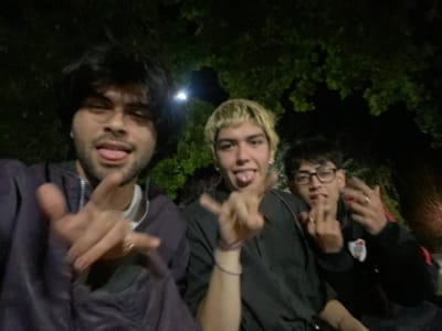 Three young men pose for an upside-down photo. The man on the left wears glasses and a black jacket with a white and red logo. The middle man has bleached blond hair and wears a dark-colored top. The man on the right wears a zip-up sweatshirt. All three stick their tongues out and make hand gestures towards the camera. They are outside in a dark area with trees in the background.