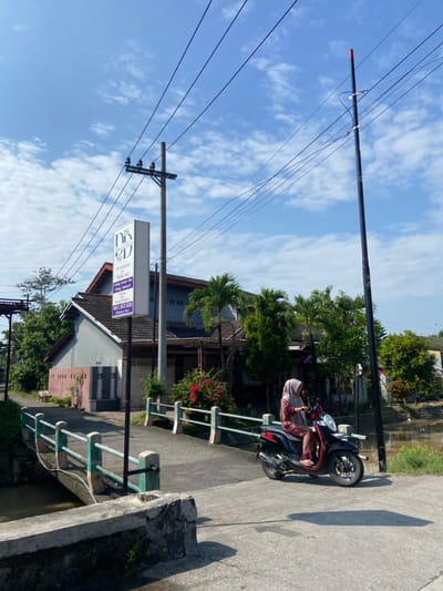 The image depicts a woman wearing a hijab riding a red and black scooter across a small concrete bridge. The bridge is flanked by a metal railing and sits over a narrow canal. The woman is traveling from left to right in the image. On the left side of the image, there is a sign advertising a business named "Dr. Liza Aesthetic Clinic". The sign is mounted on a black pole which is also supporting several electric wires running across the sky. On the left, a grey concrete building with a maroon-colored trim is partially visible. The building is set back from the street and covered in green foliage. The sky is a clear blue with white clouds, and the day is sunny.  The location is Kecamatan Tulangan, Indonesia (latitude: -7.476, longitude: 112.616).