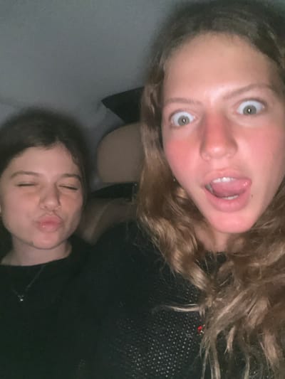 Two young females are seated in a dimly lit interior, likely a vehicle. The female in the foreground on the right has long, wavy brown hair, wide-set eyes opened wide, and her tongue extended from her open mouth, creating an exaggerated expression. Her cheeks and nose show a red flush. She is wearing a dark, possibly black, textured long-sleeved top, with a small red object visible near her right shoulder. The female on the left, partially obscured in the background, has darker, shorter hair, her eyes closed, and her lips pursed in a kissing gesture. She wears a nose ring and a thin silver necklace with a small pendant over a dark top. The background is indistinct and dark, with hints of light-colored upholstery or headrests.
Ben-Gurion Airport (TLV) is located in the Central District of Israel, between the cities of Lod and Airport City. It is situated approximately 20 kilometers (12 miles) southeast of Tel Aviv and 45 kilometers (28 miles) northwest of Jerusalem.