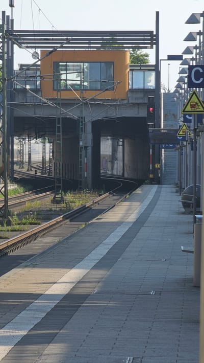 The image depicts a train station platform in Neu-Ulm, Germany, likely during the daytime. The platform is paved with concrete tiles and features tactile paving for visually impaired individuals. To the left of the platform are railway tracks with overhead catenary lines for electric trains. In the background, a concrete structure supports an elevated building with orange-colored walls and large windows, serving as part of the station's infrastructure, possibly a signal box or control center. Various signs, including station indicators and warnings, are visible along the platform. A red traffic signal is also present. The scene is devoid of people. The environment appears to be urban, with the presence of railway infrastructure and city elements.
