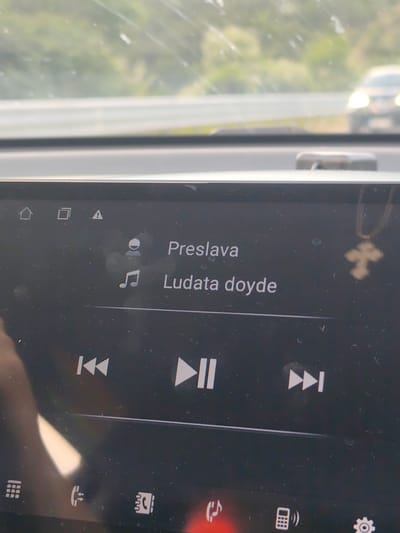 The image shows a car's infotainment screen displaying the song "Ludata doyde" by Preslava. The screen also shows various icons including home, gallery, warning, contact, phone, and settings. There is a reflection of a cross pendant in the upper right corner of the screen. The image was taken from inside a car, as the windshield and road are visible in the background, including a distant car. There is no indication that the image was taken in Jeleznitsa, Bulgaria.