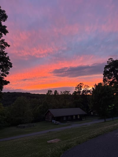 This is a horizontal, eye-level, medium shot of a scenic sunset over a wooded area with a few buildings. The sky is filled with vibrant streaks of pink, orange, and purple clouds. A silhouette of trees lines the horizon, with a dark forested area behind them. In the foreground, there are two small, rustic cabins situated on a grassy lawn. One cabin, larger than the other, has a dark roof and a few windows visible. A picnic table sits in front of it. A paved path runs through the scene, and a small, fenced-in area is visible to the left of the larger cabin. The overall impression is peaceful and natural.
