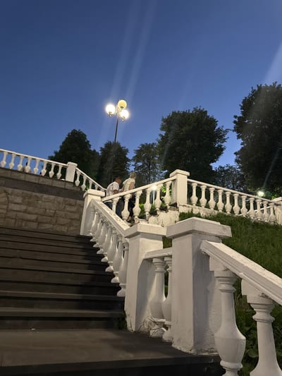 The image depicts a wide stone staircase leading up a hill at dusk. The staircase is bordered by ornate white balustrades and a stone retaining wall on the left. At the top of the stairs, a streetlamp with three globes illuminates the scene. Two people, a man and a woman, are ascending the stairs, with the man in front of the woman. They appear to be walking away from the camera. Lush green grass and trees line the upper parts of the staircase and the surrounding area. The sky is a deep blue, indicative of the twilight hour. The overall scene suggests a public park or promenade in Endirei, Russia, at the end of the day.