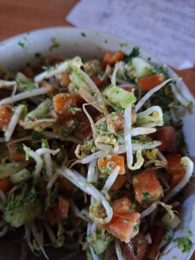The image shows a close-up of a salad in a bowl. The salad contains diced orange-red produce (likely tomatoes or carrots), light green lettuce or another leafy vegetable, and white sprouts. A green sauce or dressing coats the vegetables. A partial view of a table or surface is visible at the bottom, and a blurred piece of paper is in the upper left corner of the image. The image was taken indoors in Lodtunduh, Indonesia.