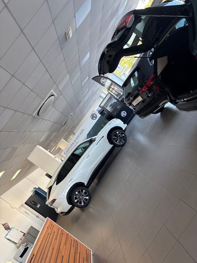 This image captures the interior of a modern car dealership showroom in Ancona, Italy, during daytime. The scene is dominated by two MG SUVs displayed on a clean, light-colored tiled floor.

In the foreground, a black MG ZS SUV is prominently featured with its trunk open, showcasing its interior. Its rear license plate, though a display plate, clearly reads "MG ZS." Further back and to the right, a white MG SUV, likely another MG model, is also on display. The showroom itself boasts a contemporary design with matching light-colored tiled floors and ceilings, featuring integrated lighting fixtures and ventilation grilles. Large windows along the left side of the showroom reflect outdoor trees and sky, indicating natural light entering the space.

Various display stands and signage are visible throughout the showroom. A prominent white stand displays the "MG" logo and the Italian text "Elettriche e non solo. della MG." (Electric and not only. of MG.), highlighting the brand's offerings. Another sign, partially visible, reads "Benvenuti a bordo." (Welcome aboard.). In the background, a black display stand features the "smart" logo (appearing inverted due to its perspective in the image). To the right, a stylish wooden-slatted counter or table adds a touch of natural texture to the otherwise sleek environment. The overall setting suggests a professional and inviting atmosphere for car shoppers.