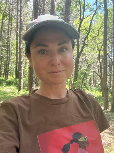 A woman is taking a selfie in a forest. She is wearing a brown t-shirt with a graphic print of a woman with sunglasses on it, and a baseball cap. She has light skin, dark brown hair pulled back, and green eyes. She is smiling slightly and looking at the camera. The background is a dense forest with tall, slender trees and lush green foliage. The lighting suggests it is daytime with the sun filtering through the trees. The location is identified as Žlijebi, Montenegro.