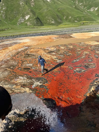 The image shows a person standing on a ledge with a dramatic drop. The person is wearing a blue shirt and grey pants, with a dark hat. They are looking out at the expansive landscape, their shadow cast long on the rocky, colorful ground. The ground itself is a striking mosaic of reds, oranges, and browns, patterned with darker veins and lighter patches, suggesting mineral deposits or dried mud. To the left, lush green hills roll into the distance, with a faint path or road cutting through them. The sky is a bright blue, indicating a clear day. Water cascades over the edge of the ledge in the foreground, creating a dynamic spray of white. The overall impression is one of awe and exploration, with the vastness of nature contrasting with the solitary figure. The scene appears to be a natural hot spring or geyser area, given the mineral-rich ground and the water flow. The image is oriented vertically.