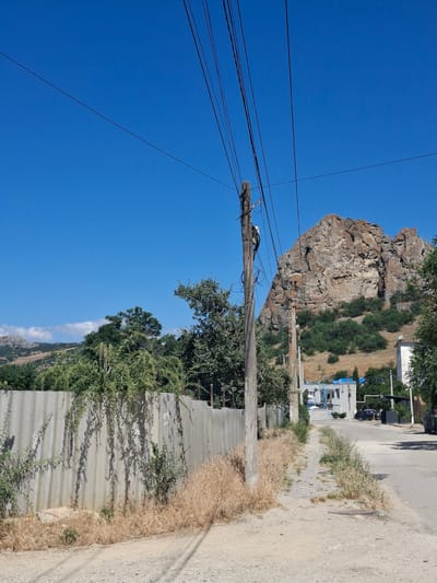 The image depicts a rural street scene in the Щебетовский поселковый совет, Ukraine, on a clear, sunny day. A dirt road runs diagonally from the foreground towards the background, with a narrow, paved pedestrian path running alongside it. A gray, corrugated fence runs along the left side of the road, overgrown with vegetation. Beyond the fence, dense green trees are visible.
In the middle ground, a weathered wooden utility pole stands prominently, adorned with numerous black electrical wires that stretch across the frame against the bright blue sky. A person, dressed in dark clothing, is seen working on the utility pole. Further down the road, a rocky, mountainous landscape dominates the background, with scattered green trees and bushes on its slopes. A white building with a blue roof is partially visible in the distance, along with other structures and parked vehicles. The ground is covered with dry grass and sparse vegetation. The overall impression is one of a somewhat unkempt but functional rural environment under a clear, expansive sky.