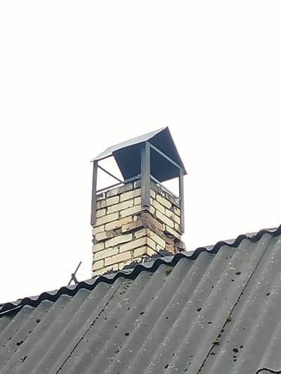 A close-up, slightly angled shot captures a brick chimney with a metal cap standing atop a corrugated roof under an overcast sky. The chimney is constructed from light-colored bricks, some of which appear weathered or damaged. The cap is a dark metal structure with a pitched roof, supported by four vertical posts. The roof itself is dark gray and shows signs of age, with some green moss growth visible in the crevices between the corrugations. The background is a uniform, pale gray, indicating a sky with low cloud cover. No people or specific landmarks identifiable with Дзешчанскі сельскі Савет, Belarus are present in the image.