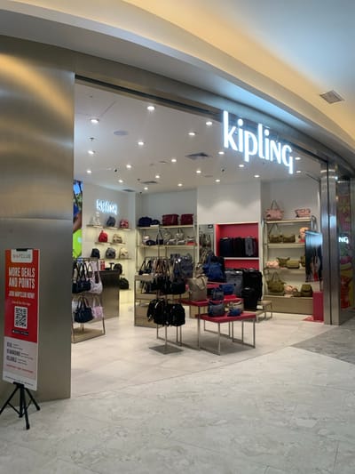 The image displays the brightly lit interior of a Kipling store, likely situated within a shopping mall in Samarinda, Indonesia. The store features an array of Kipling brand bags, including handbags, backpacks, and luggage, neatly arranged on various shelves and display tables. The interior walls are white, with some display sections highlighted by vibrant red back panels.

The store's name, "kipling," is prominently displayed in large, illuminated white letters above the entrance, with another smaller "kipling" sign visible inside. The ceiling is equipped with numerous recessed spotlights and linear light fixtures, providing ample illumination. The floor is covered with light-colored, possibly white or light gray, patterned tiles. Architectural elements like a curved ceiling and polished metallic wall panels suggest a modern retail environment. No individuals are present in the store.

In the foreground, a red promotional sign is visible, advertising "MAPCLUB." The sign encourages customers to "JOIN MAPCLUB NOW!" for "MORE DEALS AND POINTS," with instructions to "Scan & Get Our App." Other text on the sign includes "REAL," "REWARDING," "RELIABLE," "Trusted Brands," and "SHOPPING FOR EVERYONE." Two QR codes are present for downloading the app from Google Play and the App Store.