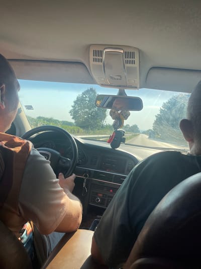 The image shows the interior of a car from the perspective of a passenger. A driver, wearing a patterned shirt, is visible from the shoulder up, operating the steering wheel. A second person is seated in the passenger seat, also seen from the shoulder up, wearing a dark green shirt. The car is driving on a road surrounded by trees and vegetation, under a clear sky. The dashboard and a rearview mirror with hanging decorations are also visible. The location is identified as Yasen, Bulgaria.
