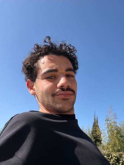 An image taken in Mougins, France, features a young adult male positioned against a clear, bright blue sky. The subject, viewed from a low angle, occupies the central and lower portions of the frame. He has dark, curly hair, a prominent mustache, and light stubble. He is wearing a dark, long-sleeved garment, possibly a sweatshirt or turtleneck, with a visible ribbed collar. His eyes are directed towards the camera, and his lips are slightly pursed. In the background, to the lower right, green foliage and the slender, pointed silhouette of a cypress tree are visible, indicating an outdoor environment under direct sunlight.