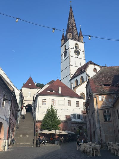 The image shows a street scene in Sibiu, Romania, featuring the Evangelical Cathedral in the background. The cathedral has a tall, white tower with clock faces and a decorative roof. In the foreground, a cobblestone street leads up a set of steps, with people walking up or sitting on them. Buildings line both sides of the street, some with red tile roofs and others with white facades. A restaurant or cafe with outdoor seating is visible on the right side of the street, with patrons seated at tables under umbrellas. Above, string lights stretch across the street. The sky is clear and blue.