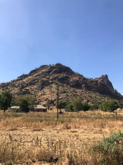 This image captures a rural landscape in Bundot, Nigeria, under a clear, bright blue sky. A prominent, large rocky hill, characterized by exposed dark rock formations and sparse dry vegetation, dominates the background. Scattered green trees are visible on the lower slopes of the hill, which also features a distinct, sharply pointed rock formation on its right side. Multiple utility lines span across the mid-section of the hill.
In the midground, a paved road extends horizontally across the frame. A dark utility pole with several overhead wires stands on the left side of the road. Adjacent to the road on the left, two residential-style buildings are visible: one light green with a light-colored roof, and another red with a similarly colored roof. To the right of the utility pole, a partially collapsed or unfinished light-colored structure is present. Various trees with green foliage and some dry, leafless trees are interspersed throughout this area. A single individual is observed operating a motorcycle on the road, traveling from left to right. Further along the road to the right, two indistinct figures are partially visible near a small, light-colored structure.
The foreground consists of a field covered primarily with dry, yellow-brown grass and scattered patches of green shrubbery.