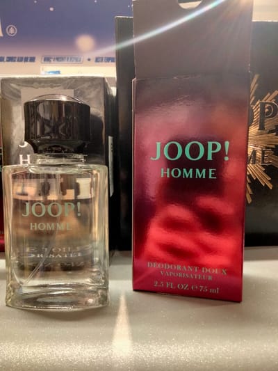 A bottle of Joop! Homme cologne stands next to its packaging. The clear bottle, with a black cap, displays the brand name "Joop! Homme" and "E TOILE DR SATEE." The packaging is a glossy red and displays "Joop! Homme" in green text and "DEODORANT DOUX VAPORISATEUR 2.5 FL OZ e 75 ml". Behind, more products are visible, adding depth to the shot. The surface underneath reflects light.