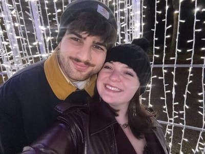 The image captures a happy couple, a man and a woman, posing closely together in what appears to be a festive outdoor setting at night in Arezzo, Italy. Both individuals are looking towards the camera with bright smiles.
The woman, positioned on the left, has dark hair and wears a dark jacket, possibly leather, and a black knitted beanie with a pom-pom. A ring is visible on her left hand. The man, on the right, has dark hair, a mustache, and a short beard. He is dressed in a dark jacket with a prominent mustard-colored collar and what appears to be a light-colored shirt underneath. He wears a dark beanie on his head, which clearly displays a "Carhartt" patch.
The background is filled with numerous strings of white, glowing fairy lights arranged in vertical lines, creating a luminous, grid-like pattern against a dark, out-of-focus backdrop. This elaborate lighting suggests a decorative display, such as a Christmas market or a light festival. The warm clothing worn by both subjects indicates cool weather. The overall atmosphere conveyed by their expressions and the twinkling lights is joyful and celebratory.
