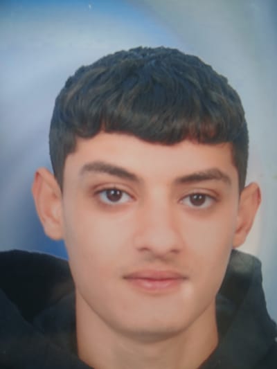 The image is a close-up, head-and-shoulders portrait of a young male with dark, short, bowl-cut hair. He has dark eyes, prominent eyebrows, and a light mustache and goatee. He is wearing a dark hooded jacket. The background is a soft, blurry gradient of light blue. The image appears to be a photograph, possibly for identification purposes, taken indoors. There is no visible indication of the city of Hun, Libya, in the image itself.