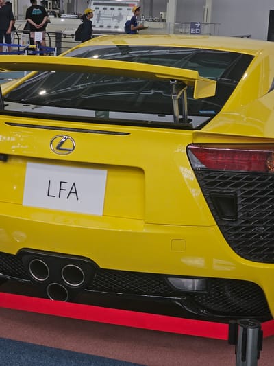 A bright yellow Lexus LFA sports car is parked indoors, likely at an automotive exhibition or dealership. The rear of the car is prominently displayed, showing its distinctive triple exhaust pipes, a large rear spoiler, and the "LFA" badge on a white plate. The car is cordoned off with a red velvet rope barrier. In the background, several people are visible, some wearing casual clothing and others in uniforms, suggesting an event setting. The environment appears to be a convention center or exhibition hall, with industrial-style flooring and overhead lighting. The automotive context of Nagoya, Japan, is implied by the presence of a Lexus, a Japanese luxury vehicle brand, and the overall presentation of a sophisticated automobile.