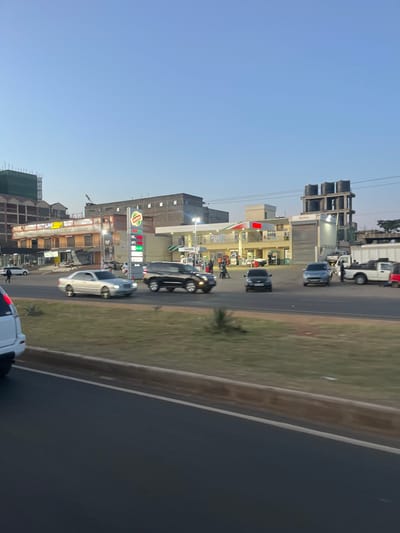 The scene depicts a late afternoon or early evening urban environment in Biashara ward, Kenya, captured from a vehicle on a multi-lane roadway. In the immediate foreground, a white SUV's rear bumper and wheel are partially visible on the left. The road surface is dark asphalt, bordered by a light brown concrete curb, beyond which lies a grassy median strip with sparse dry vegetation and small shrubs.

In the midground, vehicles are in motion on the road parallel to the median. A silver Mercedes-Benz sedan proceeds left-to-right, followed by a black SUV, also moving left-to-right. A dark-colored sedan or small SUV faces the camera, positioned on the road's far side. To the right, a prominent Shell petrol station features multiple fuel pumps beneath an illuminated canopy. A tall vertical sign displays the Shell logo, "Burger King" branding above, and lit fuel prices for V-Power Diesel (red), Shell FuelSave Diesel (green), and Shell FuelSave Unleaded (blue). Approximately 5-7 individuals are visible at the petrol station, some appearing to be staff in light-colored uniforms, others possibly customers, engaged in walking or standing near the pumps. The petrol station complex includes a multi-story building with a facade featuring green, white, and red stripes. Adjacent to the petrol station on the right, another commercial building has a large roll-down shutter and, further right, a scaffolding structure supports several large black water tanks on its upper levels. A light-colored pickup truck and another vehicle are parked or moving slowly near this area.

In the background, a row of commercial buildings is visible. On the far left, a multi-story building is partially obscured by green construction netting and scaffolding. Other buildings are multi-story structures, predominantly of masonry construction. A construction crane is faintly discernible between two buildings in the distance on the left. The sky above is a clear, light blue, indicative of dusk or dawn.