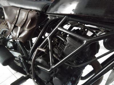 A close-up, angled forensic image depicts the right side of a black motorcycle, likely situated indoors on a light-colored floor in Majdalena, Czechia. The motorcycle features a dark brown leather saddlebag secured below a black textured seat, displaying two vertical straps with brass-colored rivets and buckles. The exposed engine, primarily black with visible cooling fins and a partially obscured "V HON" emblem on a silver-colored cover, is encased by a prominent black tubular frame. A polished or chrome U-shaped bar is integrated into the frame, partially shielding the engine. A black, cylindrical exhaust muffler extends horizontally from the lower left side of the frame. A black rubberized footpeg is positioned below the engine. A knobby rear tire is partially visible behind the saddlebag, suggesting an off-road or dual-sport capability. The background is out of focus, showing a light-colored surface. No individuals or discernible actions are present.