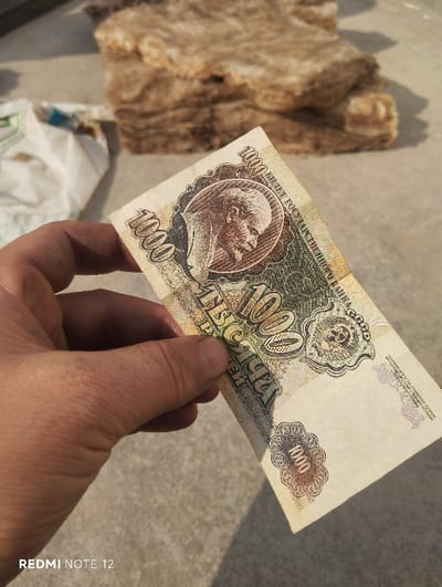 A light-skinned adult hand, showing some surface dirt, holds a 1000 Soviet Ruble banknote (1991 series) in the foreground. The banknote displays a portrait of Vladimir Lenin on its left side and the denomination "1000" in Cyrillic. The banknote appears slightly creased and worn. The scene is outdoors, brightly lit by direct sunlight, casting shadows from the hand onto the surface below. The immediate surface beneath the hand and banknote is a light grey, textured material, possibly concrete or paving. In the mid-ground, a stack of irregular, fibrous, light brown material, resembling insulation or rock wool, is visible. To the far left, a crumpled white plastic bag with green markings is partially present. The specific location in the city of Qizilsay, Uzbekistan, cannot be determined from the visual information provided in the image.