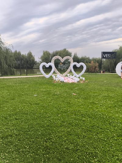 This image captures an outdoor wedding proposal setup in Komsomolskoe, Kyrgyzstan. The focal point is a large, heart-shaped arch adorned with white floral decorations, with three individual heart shapes in varying sizes forming the centerpiece. A smaller, central heart has the text "Will you Marry me?" inscribed on it. The area around the arch is decorated with scattered rose petals and small white floral accents, suggesting a romantic proposal.

The setting is a lush green lawn dotted with small white flowers, indicative of a park or outdoor venue. In the background, a tranquil body of water, likely a lake or river, is visible, bordered by a line of trees. The sky is overcast with heavy clouds, suggesting a late afternoon or early evening time of day.

To the right of the heart arch, a black sign with the white text "VESLO" and a musical note logo is prominently displayed. Near the sign, there's a white circular prop and a person, possibly a photographer or part of the event setup, dressed in casual attire. The overall atmosphere appears to be set for a special occasion, likely a marriage proposal, with a romantic and serene ambiance. The weather appears to be cloudy but not actively raining.