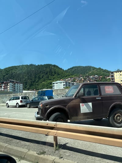 The image depicts an outdoor scene, likely in Zvornik, Bosnia and Herzegovina, with a clear blue sky and a verdant, tree-covered hillside forming the backdrop. The hillside is dotted with houses. In the foreground, a brown, older model SUV is parked on a paved surface, partially obscured by a yellow metal guardrail. The SUV has a white sticker with "mtel" in red letters on its rear passenger window. Another white rectangular sticker with text in a foreign language and "mtel a.d." is visible on the side of the vehicle. To the left of the SUV, a dark gray sedan and a white minivan are also parked. Behind the vehicles, a concrete wall with graffiti is present, and beyond that, several multi-story buildings are visible. The overall impression is of a daytime scene in a semi-urban or suburban environment.