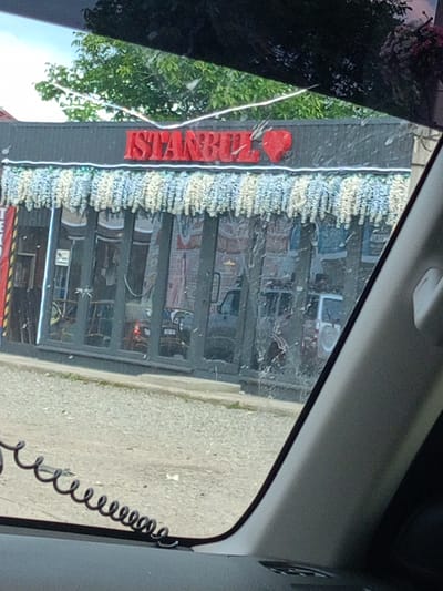 The image shows a street-level view of a building with a red sign that reads "ISTANBUL" followed by a red heart. The building has a dark-colored facade with large glass windows. A decorative string of white and blue tassels hangs above the windows. The windows reflect the environment, showing what appear to be vehicles and signage inside. The area in front of the building is paved with gravel. A black coiled cable is visible in the lower left corner of the image. The building is located in Zelenchukskaya, Russia, as indicated by the context.